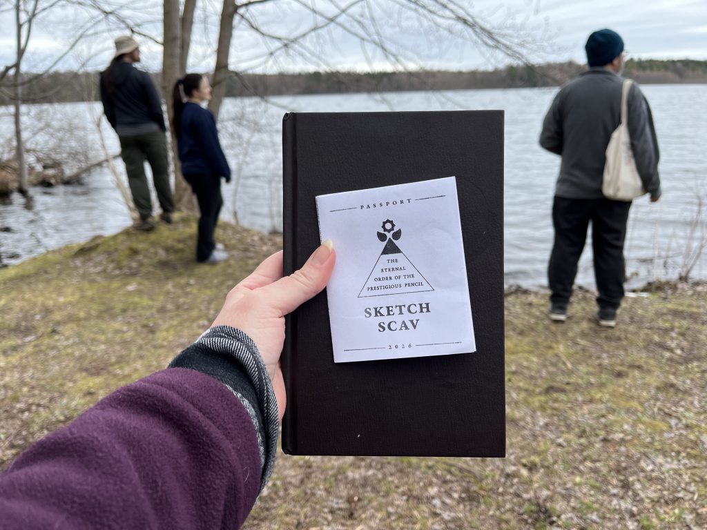 An outstretched arm and hand hold a sketchbook and an EOPP Sketch Scav passport zine on a backdrop of the outdoors near a lake. In the background, three people look out over the lake.